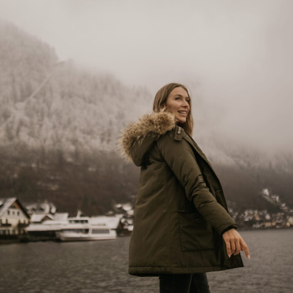 A woman wearing a green parka with a fur-lined hood smiles while standing by the lake in Hallstatt, Austria, during winter. Snow-covered houses and misty, tree-lined mountains create a serene, picturesque backdrop.